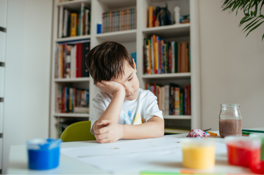 Young boy sitting at a table with his head resting on his hand, appearing tired or frustrated while working on an activity.